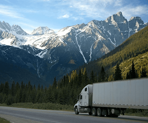 White semi truck driving on a highway with mountains and forest in the background.