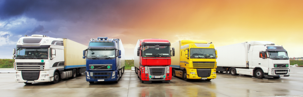 The image shows five semi-trucks parked side by side on a wet pavement with a dramatic sky in the background. The trucks are of different colors: white, blue, red, yellow, and white, arranged from left to right. The scene suggests a diverse fleet ready for Trucking Logistics, under a sky that transitions from stormy clouds to a warm sunset