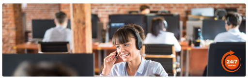 Customer service representative wearing a headset and smiling while assisting a caller in a busy call center with 24/7 service, highlighting dedicated support team in a modern office environment.