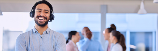 A smiling customer service representative wearing a headset stands in the foreground, with a group of colleagues engaging in a conversation in the blurred background. The setting appears to be a bright, modern office environment.