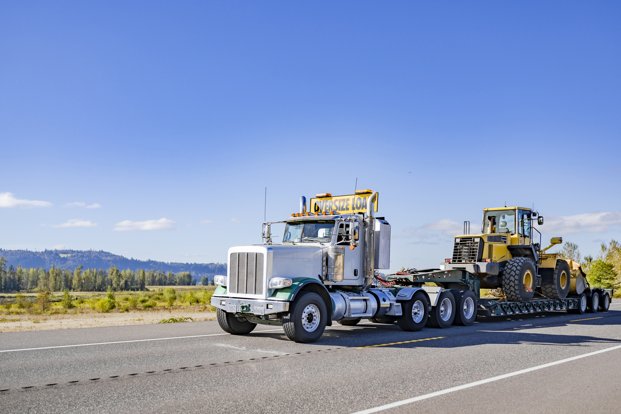 Oversize load semi-truck transporting heavy construction equipment on a lowboy trailer along a rural highway.